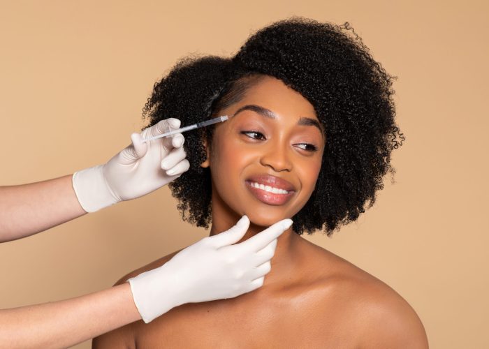 Smiling young black woman receiving professional cosmetic treatment with syringe, demonstrating beauty procedure against beige background